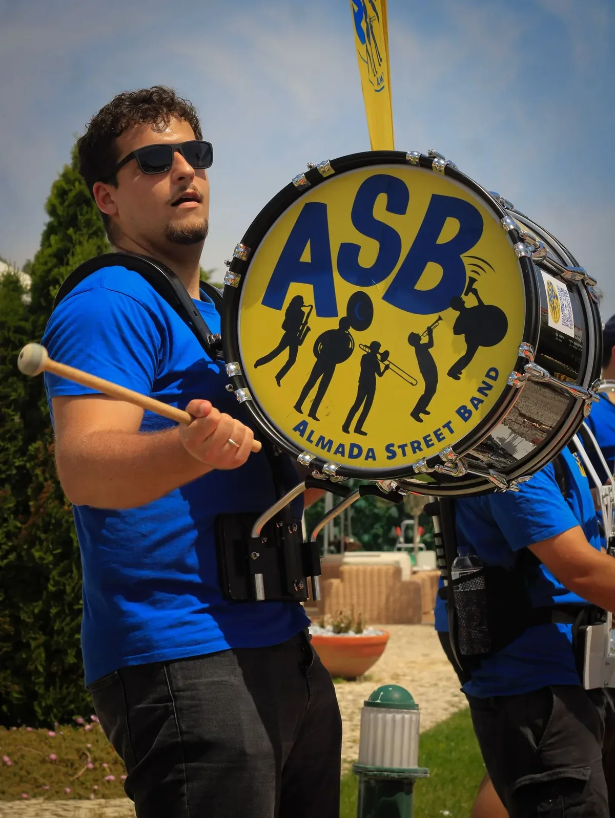 Um percussionista da banda, de óculos de sol e camisola azul, a tocar num bombo amarelo com o logótipo ASB e a silhueta dos músicos, ao ar livre.