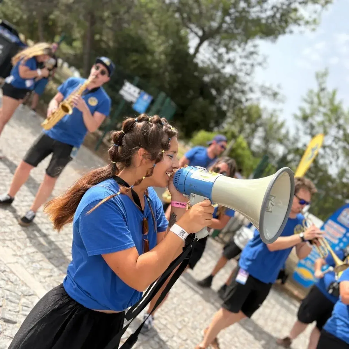 Uma música da AlmaDa Street Band, de camisola azul, a rir e a falar para um megáfone numa rua de calçada, com outros músicos da banda a tocar ao fundo.