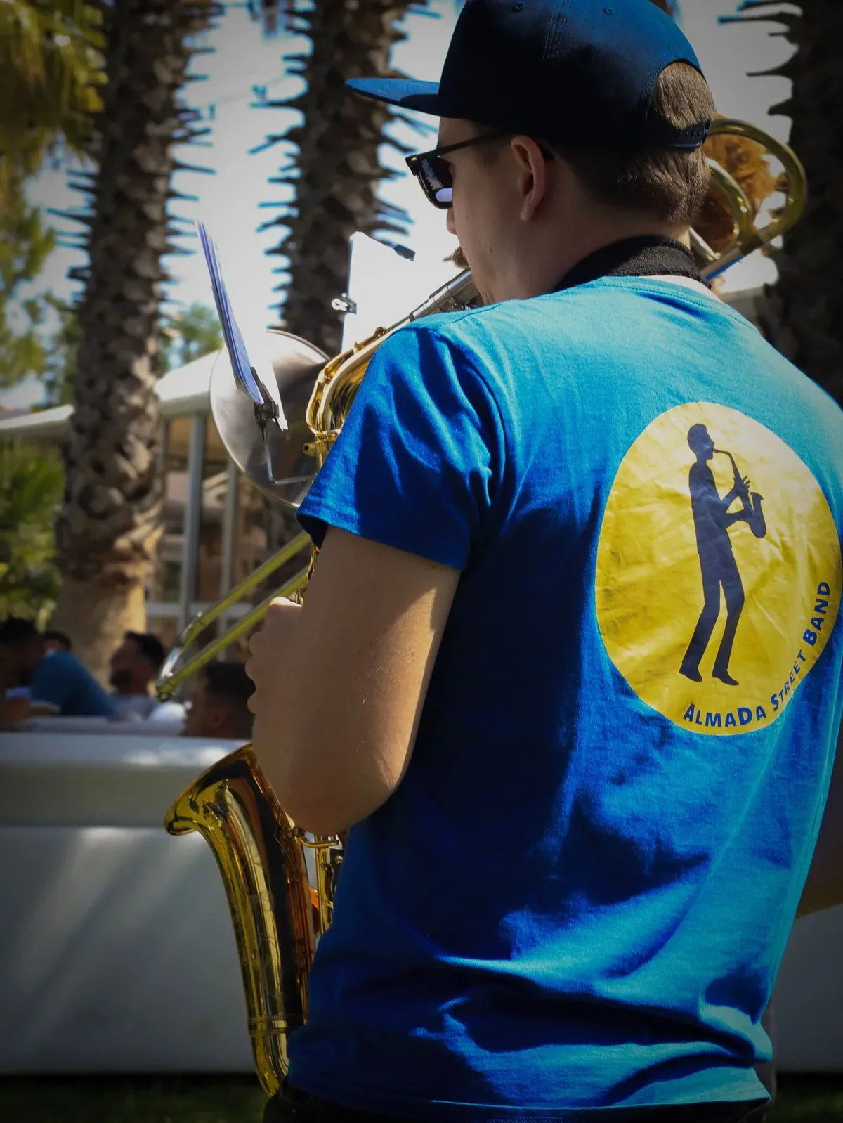Um músico de boné, visto de costas, a tocar um instrumento de metal; na camisola azul, o logótipo amarelo com a silhueta de um saxofonista e as palavras AlmaDa Street Band.