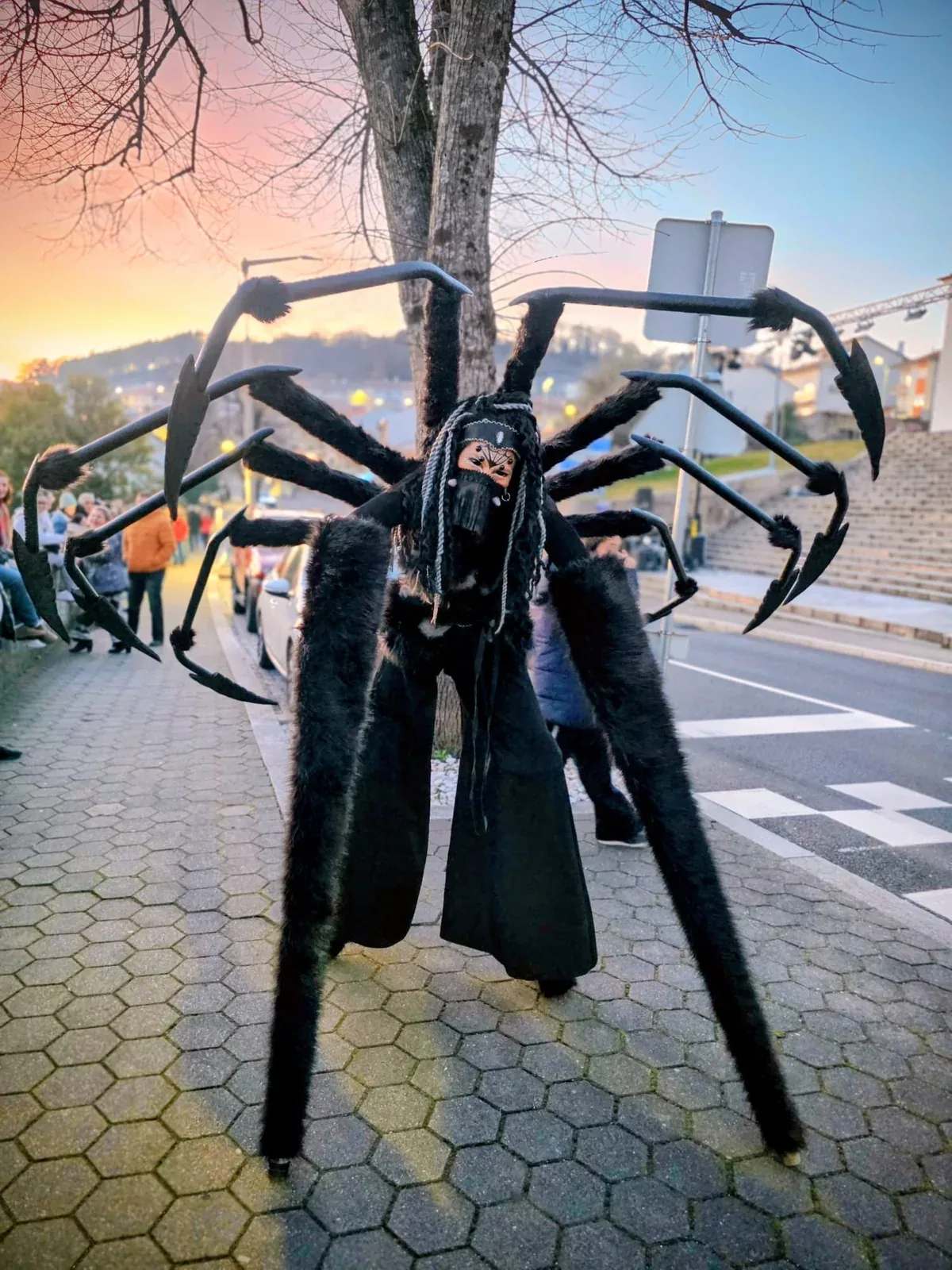 Criatura gigante em andas, com oito patas negras e máscara, a percorrer uma rua de calçada ao fim do dia.