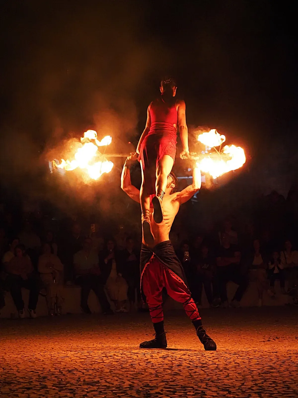 Acrobacia a dois à noite: uma artista de pé nas mãos do parceiro, com chamas nas pontas de uma barra, diante do público.