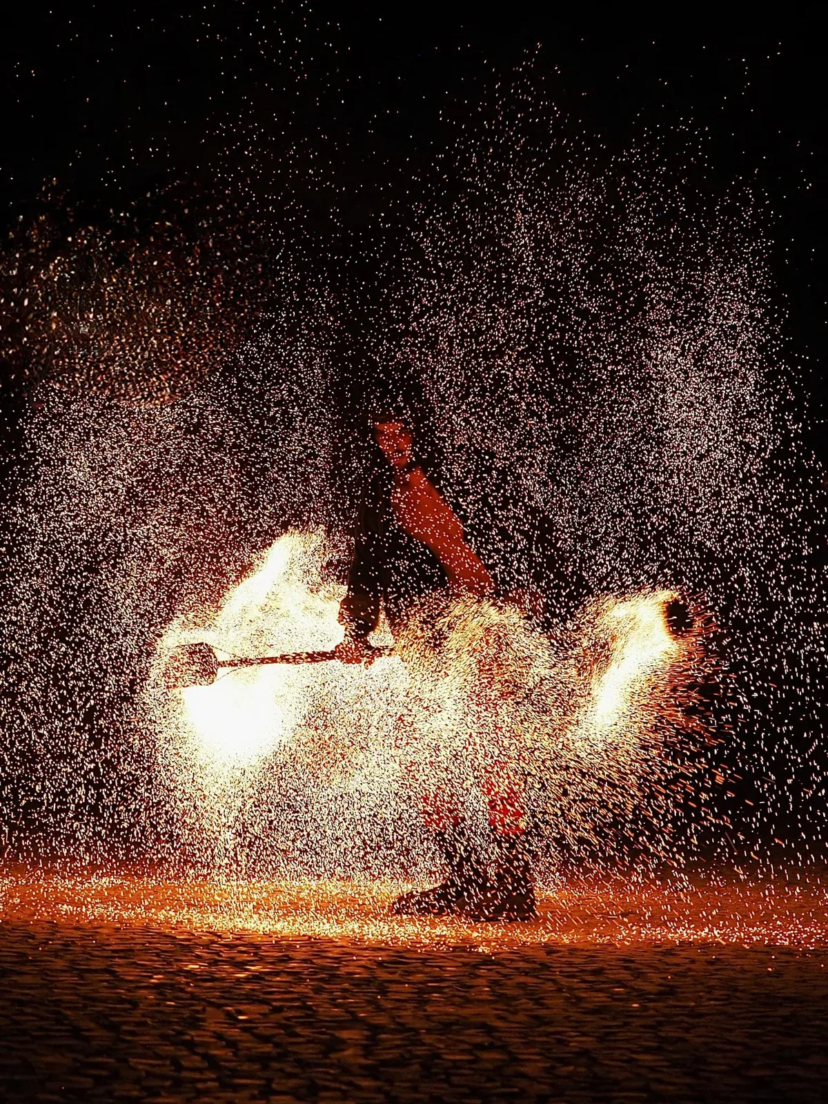 Performer a girar uma peça em chamas, envolvido numa chuva de faíscas sobre a calçada.