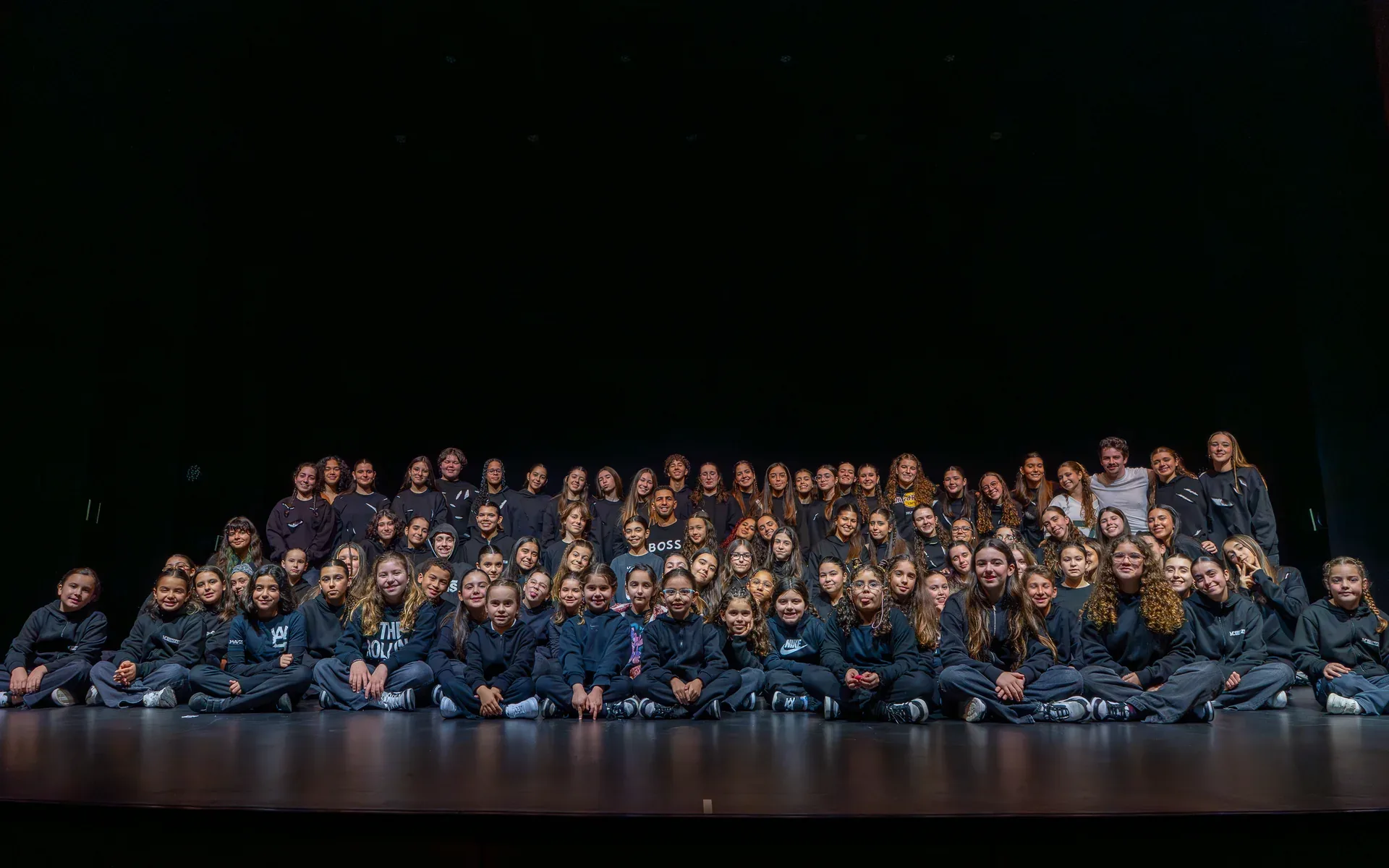 O grupo We Can Dance reunido em palco escuro, sentado e de pé, a olhar para a câmara.