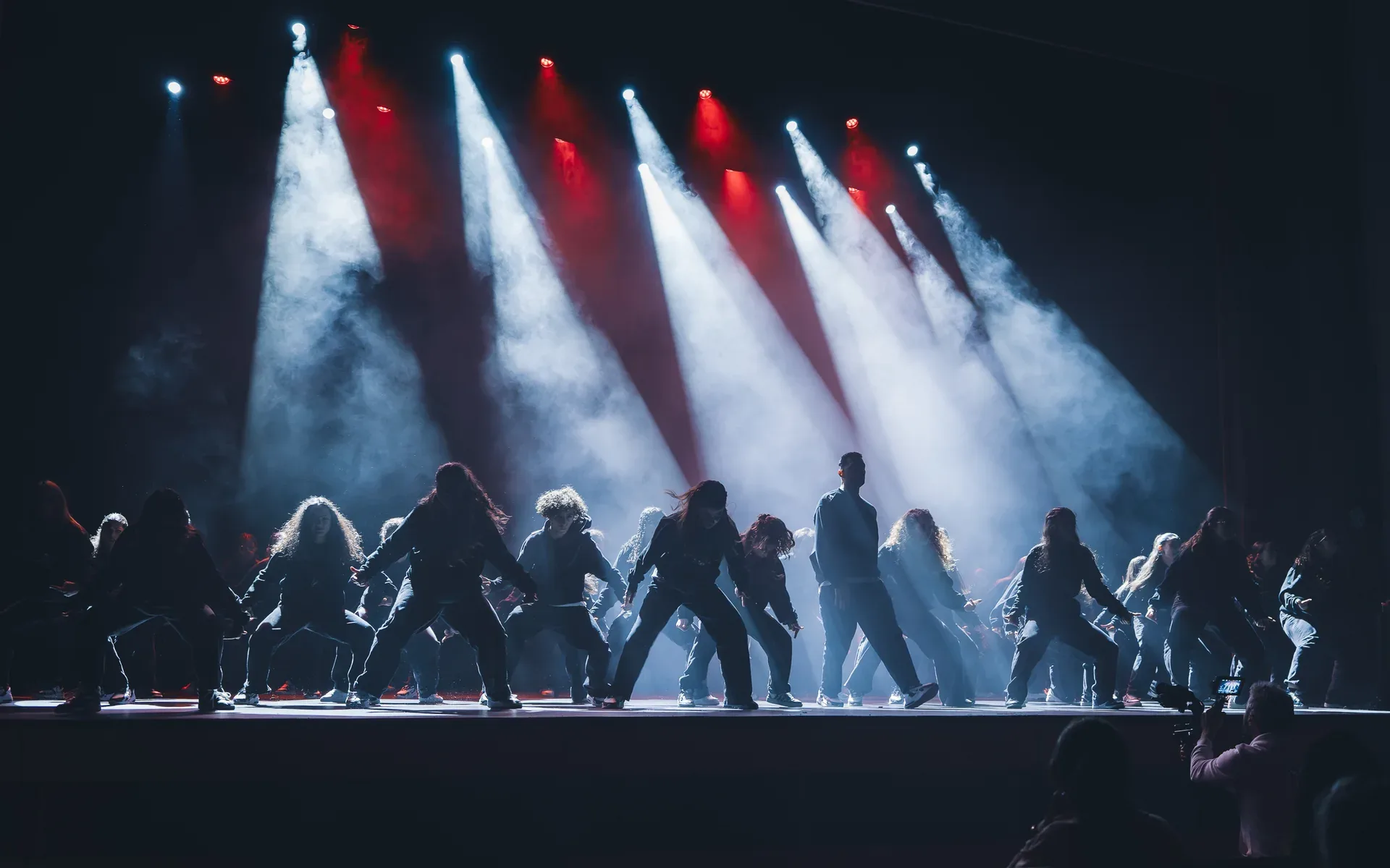 Os We Can Dance em palco: silhuetas de dançarinos sob feixes de luz branca e vermelha, entre fumo.